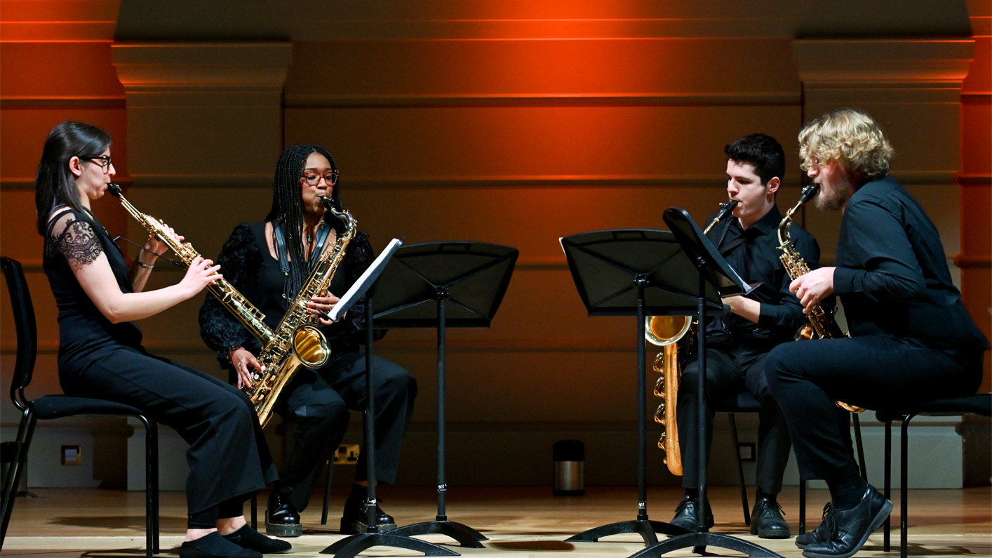 Group of students performing saxophones on stage, wearing smart black clothing.